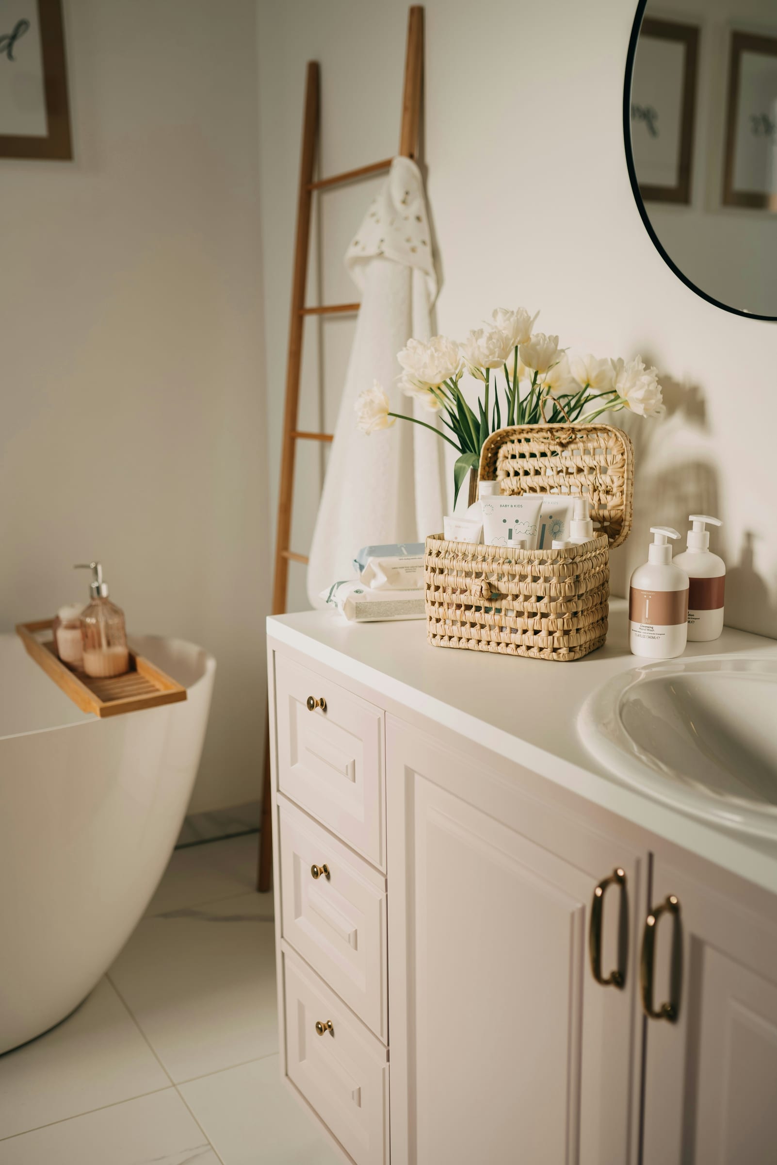 A bright, airy bathroom with a freestanding tub, wooden ladder, white tulips, and a woven basket of toiletries