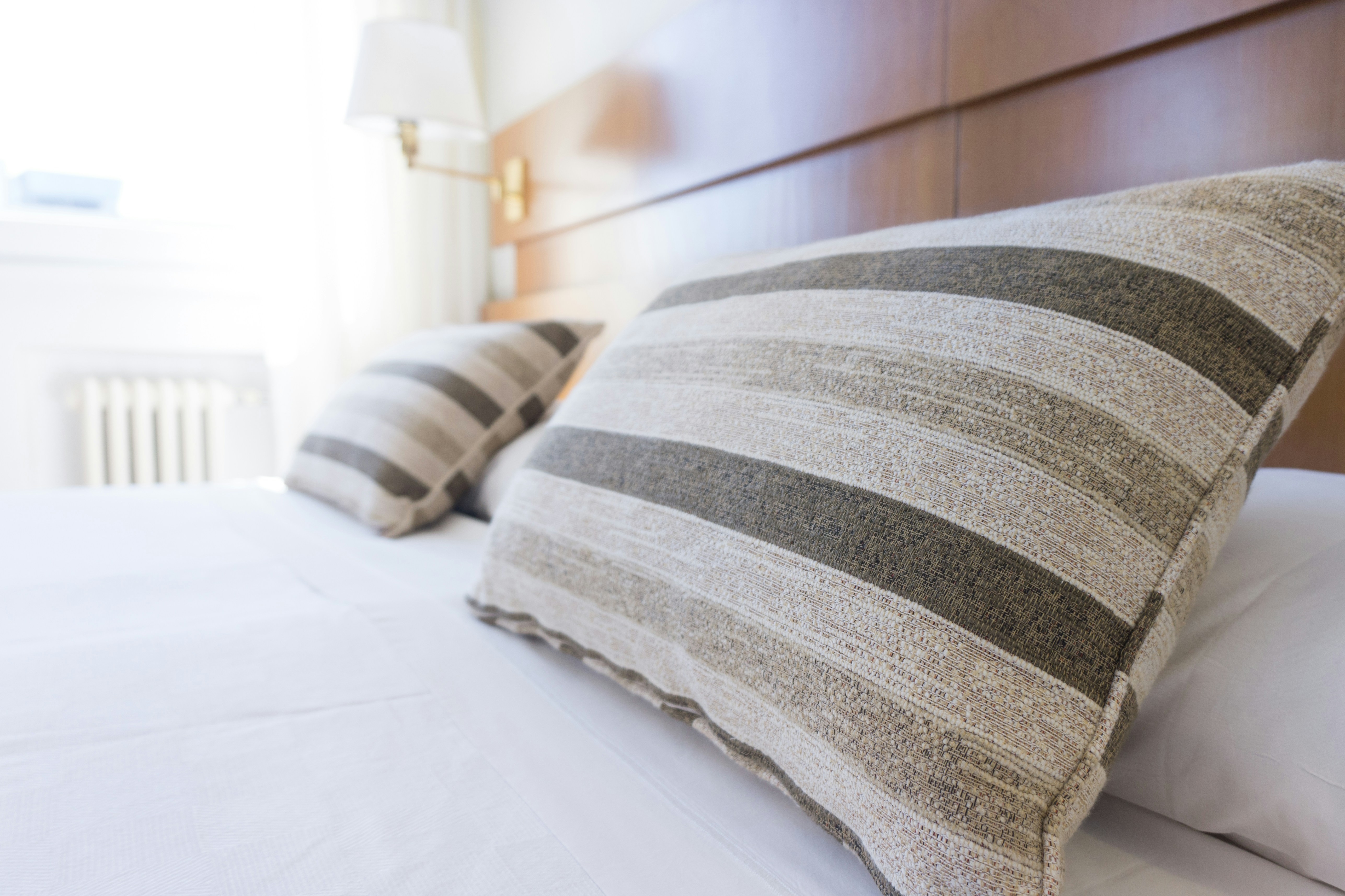 A cozy bedroom close-up with textured striped throw pillows in neutral tones on crisp white bedding against a warm wood headboard
