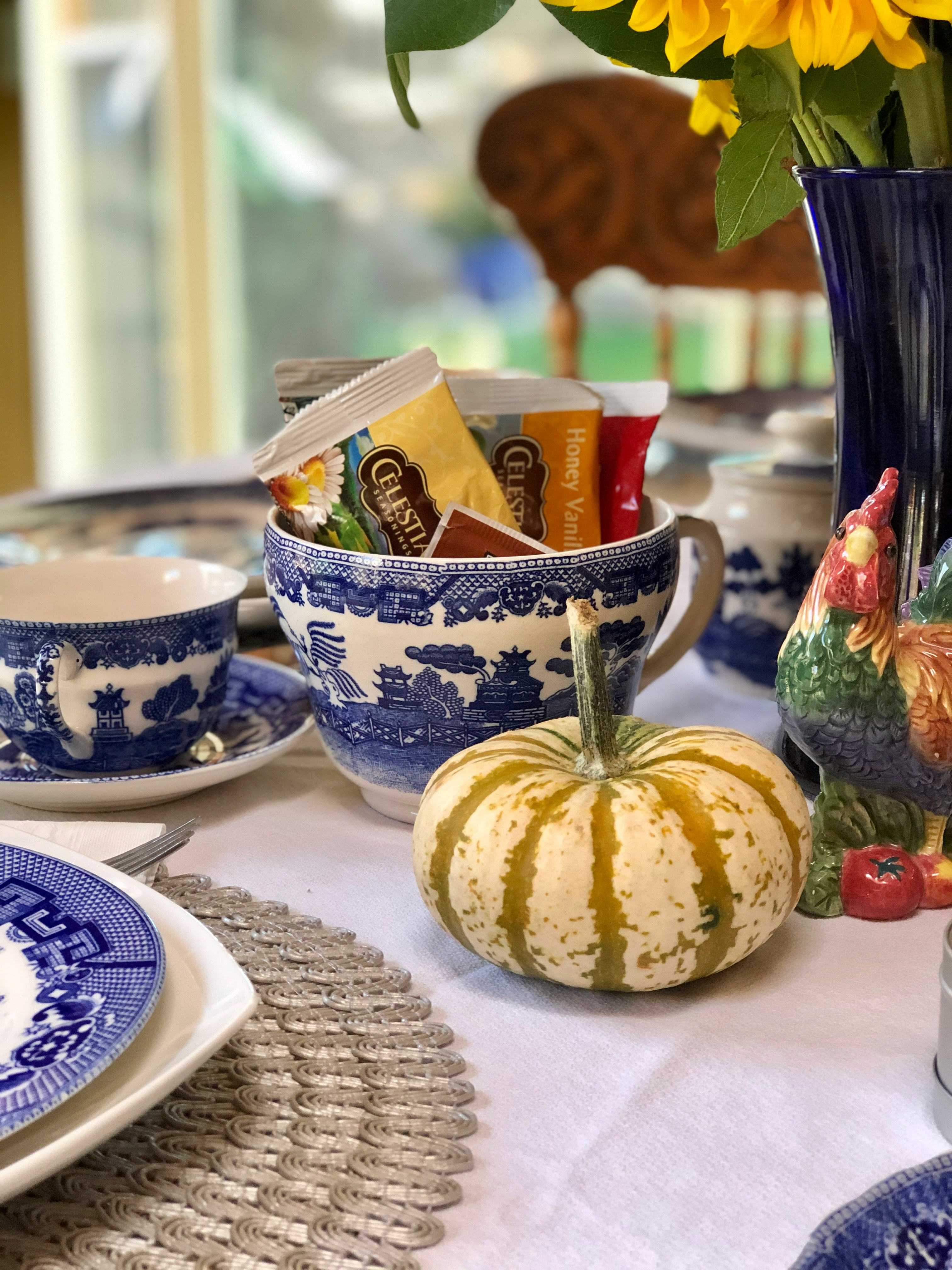 A Blue Willow china tea setting with a teacup of assorted tea bags, a small striped gourd, a ceramic rooster, and sunflowers in a cobalt vase