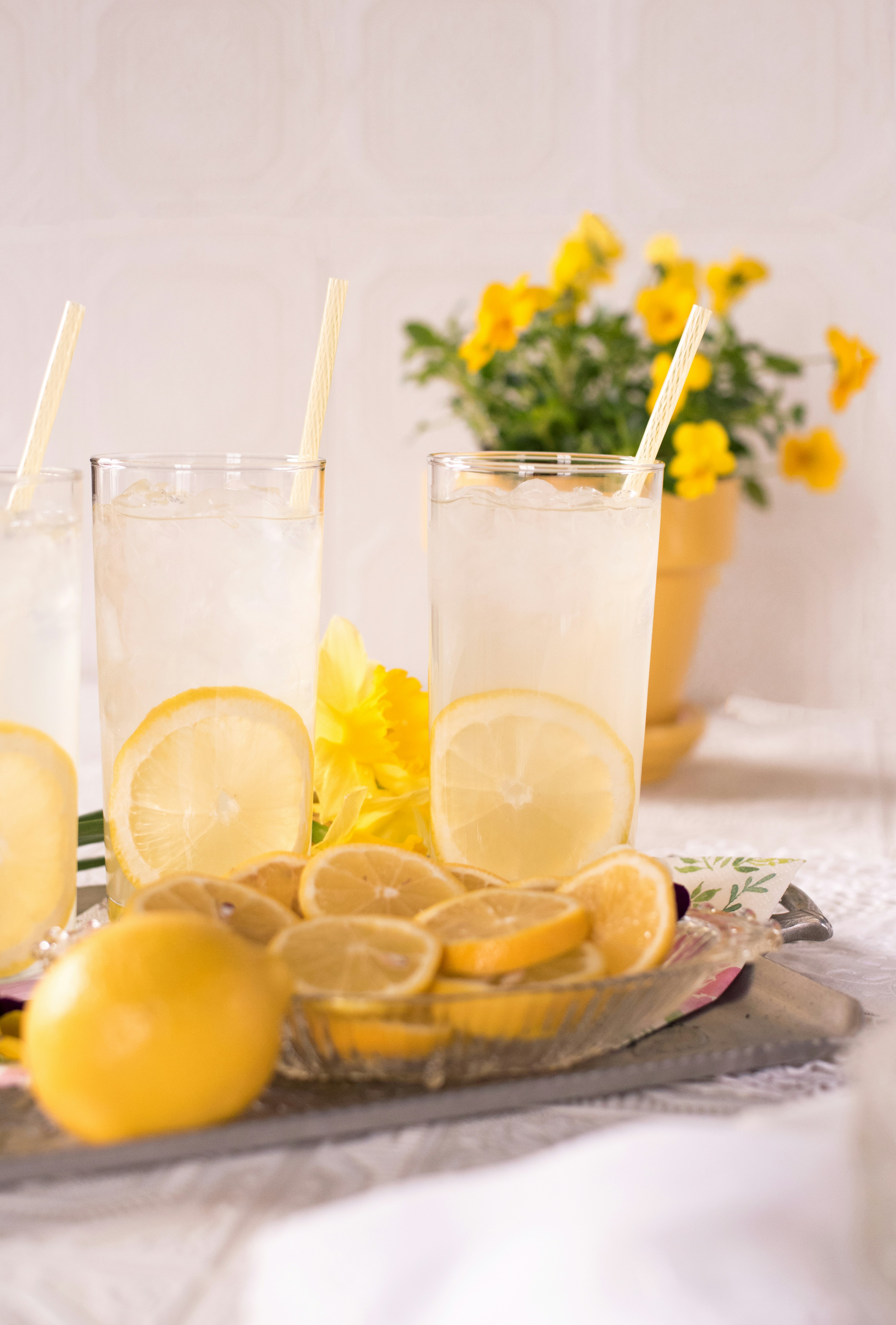 Tall glasses of iced lemonade with lemon slices and paper straws beside yellow flowers