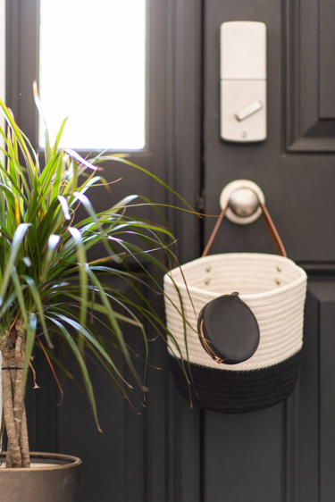 A two-tone woven rope basket with a leather strap hanging on a dark navy door, beside a leafy dracaena plant