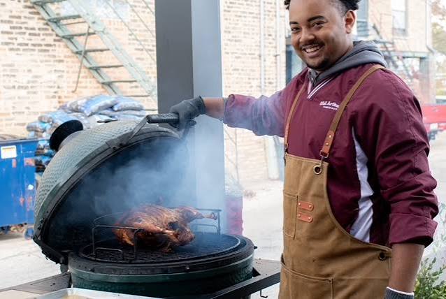 A smiling pitmaster lifting the lid of a ceramic smoker with a whole chicken cooking inside
