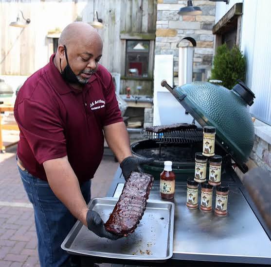 A pitmaster in an Old Arthur’s polo setting a rack of ribs on a sheet pan beside jars of Old Arthur’s sauce and rub